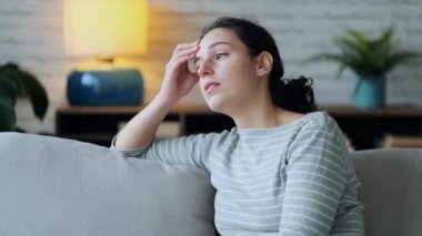 Video of worried young woman thinking while sitting on sofa in the living room at home.