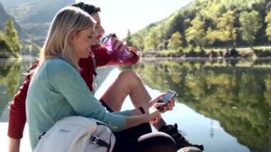 Video of two travel hikers using mobile phone and drinking water while sitting in front of the lake in mountain.