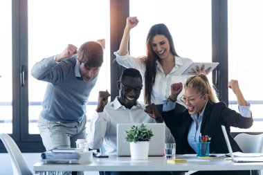 Birlikte çalışma alanında dizüstü bilgisayara bakarken bir şeyleri kutlayan başarılı genç iş adamlarından oluşan bir grup..