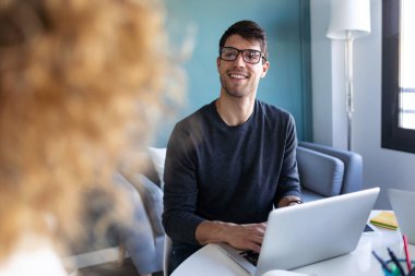 Sıradan genç girişimcinin bilgisayarını kullanırken ve yeni ofisteki meslektaşıyla konuşurken görüntüsü..