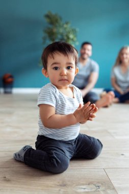 Evde yerde otururken kameraya bakan bebek portresi. Arka planda, ailesi ona bakarken oturuyor..