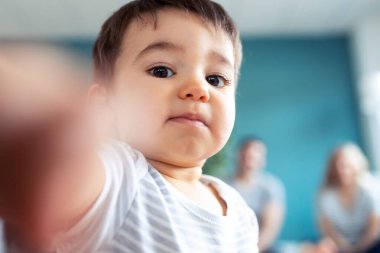Evde yerde otururken kameraya bakan bebek portresi. Arka planda, ailesi ona bakarken oturuyor..