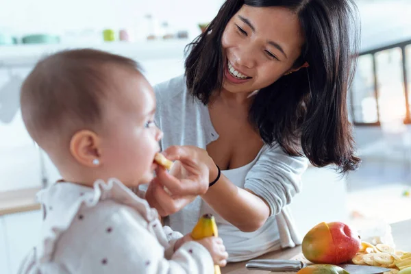 幸せな若いですお母さん給餌彼女の可愛いです赤ん坊女の子とともにバナナでザ台所にホーム — ストック写真