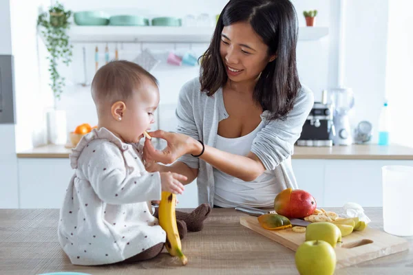 幸せな若いですお母さん給餌彼女の可愛いです赤ん坊女の子とともにバナナでザ台所にホーム — ストック写真