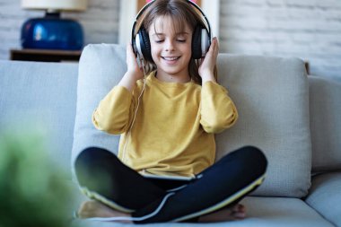Evdeki kanepede otururken kulaklıkla müzik dinleyen küçük bir kız..