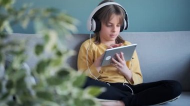 Evde kanepede otururken kulaklık ve dijital tabletle müzik dinleyen küçük kızın videosu..