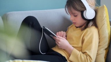 Evde kanepede otururken kulaklık ve dijital tabletle müzik dinleyen küçük kızın videosu..