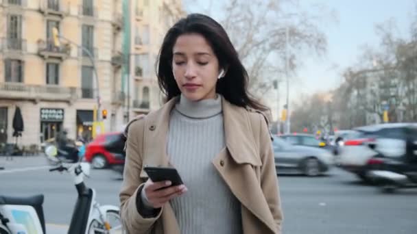 在街上散步时 漂亮的年轻女子用手机和无线耳机听音乐的视频 — 图库视频影像
