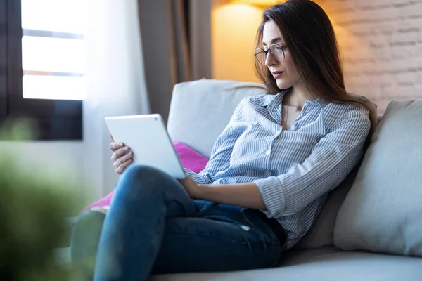 かなり若いですショットの彼女のデジタルタブレットを使用して読書のために座っている間にソファに自宅 — ストック写真