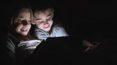 Geceleri battaniyenin altında yatıp tablet bilgisayarda video izleyen şirin bir kız ve erkek çocuğu videosu..