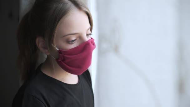 在世界范围内的病毒传播期间 悲伤的女孩戴着布面面具透过窗户看世界范围内的病毒传播 — 图库视频影像