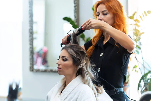 Shot of beautiful hairdresser using a curling iron to make a hair style to her woman client in a hair shop.