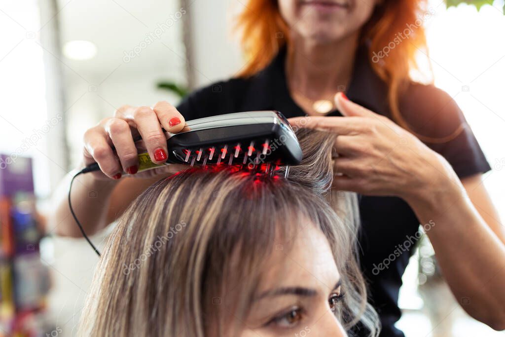 Close-up of hairdresser doing a treatment with ultrasonic and infrared laser comb for hair regrowth in her woman client at the salon.