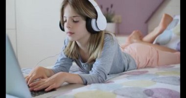 Kulaklık takan ve yatakta yatarken dizüstü bilgisayar kullanan genç bir kızın videosu. Uzaklık öğrenme kavramı.