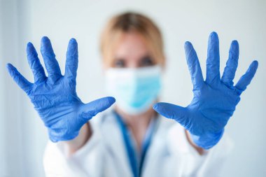 Beyaz arka planda mavi nitril eldivenler giyerken elleri kameraya gösteren tıbbi maskeli bir kadın portresi..