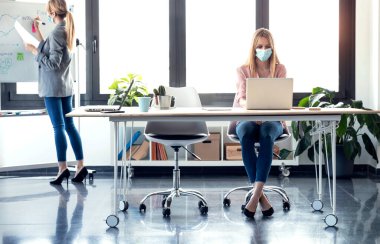 Hijyenik yüz maskesi takan genç kadınların güvenli mesafeleri korurken ofiste çalışırken çekilmiş fotoğrafları..