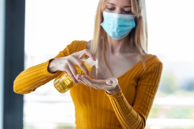 Shot of young woman with hygienic mask applying a sanitizing gel liquid for cleaning her hands. Prevention at home. clipart