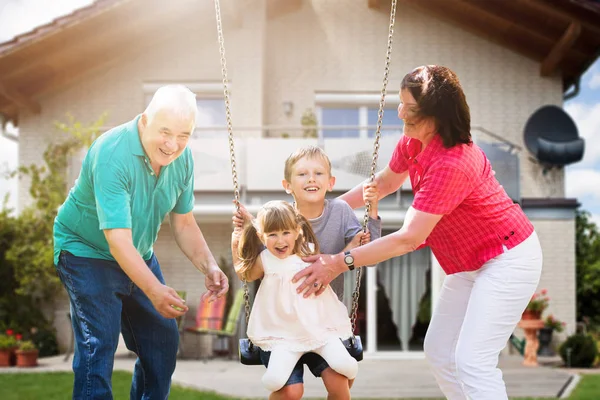 Feliz Abuelos Mirando Sus Nietos Jugando Swing Frente Casa —  Fotos de Stock