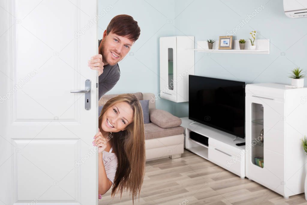 Happy Young Couple Peeking Behind Door In Living Room