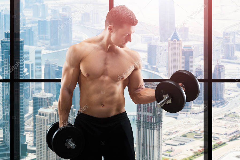 Young Muscular Man Working Out With Dumbbells In Front Of Window Overlooking The City 