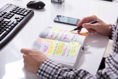 Close-up Of A Businessperson With Mobile Phone Writing Schedule In Diary On Desk