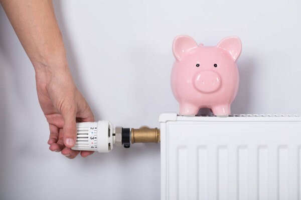 Close-up Of Man's Hand Adjusting Thermostat With Piggy Bank On Radiator Against Wall