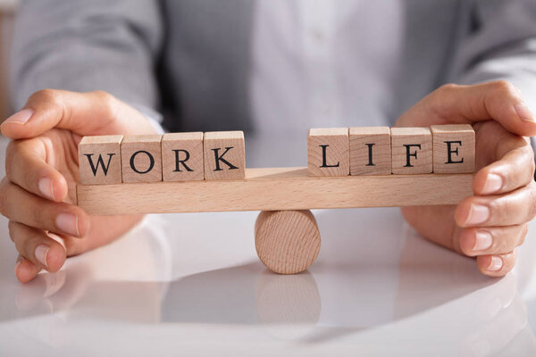 Businessperson's Hand Covering Balance Between Life And Work On Wooden Seesaw In Office