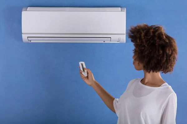 リモコン付きの青い壁に取り付け エアコンを調整する若い女性 — ストック写真