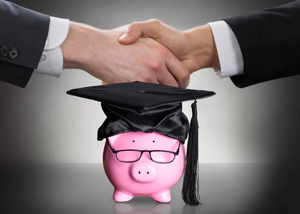 黑色毕业帽和眼镜在两个人握手前的小猪银行 — 图库照片