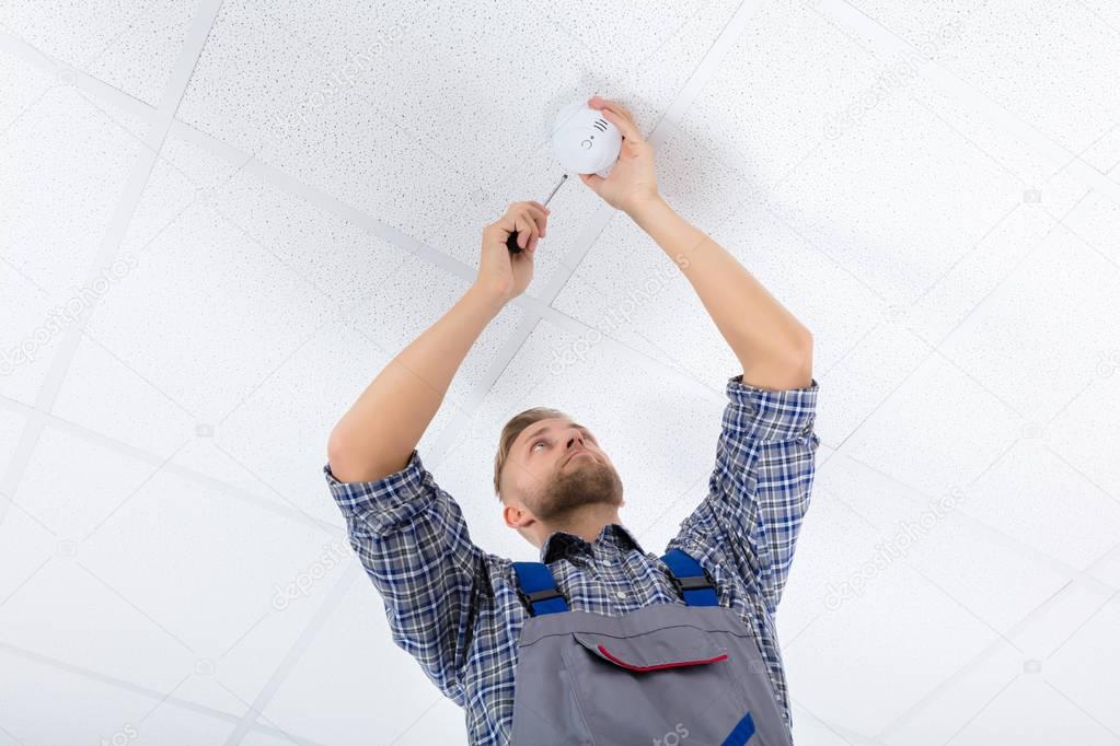Low Angle View Of A Male Electrician Fixing Smoke Detector Using Screwdriver On Ceiling Wall