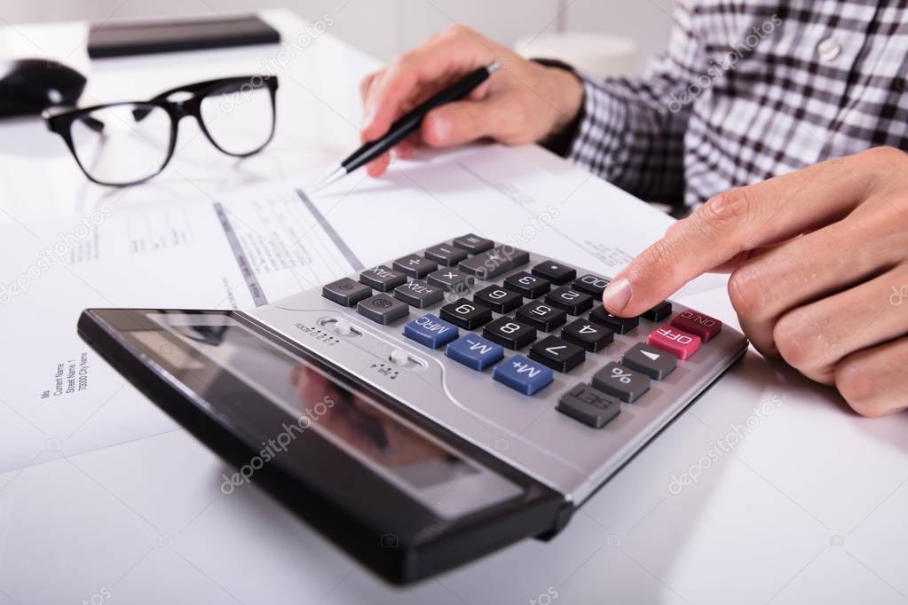 Close-up Of Businessman's Hands Calculating Invoice Using Calculator With Eyeglasses On Desk