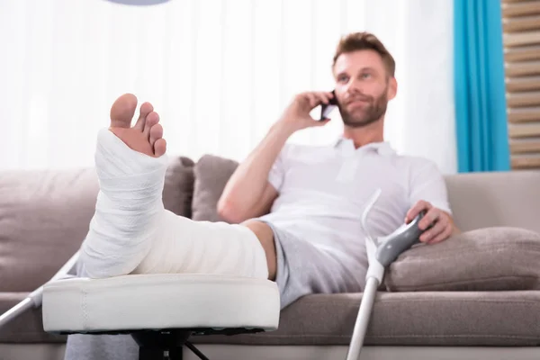Szczęśliwy Młody Mężczyzna Złamaną Nogą Siedząc Kanapie Mówić Smartphone — Zdjęcie stockowe