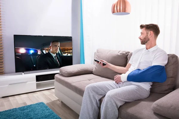 自宅のテレビで映画を見ながらソファに座って手を骨折した若い男 — ストック写真