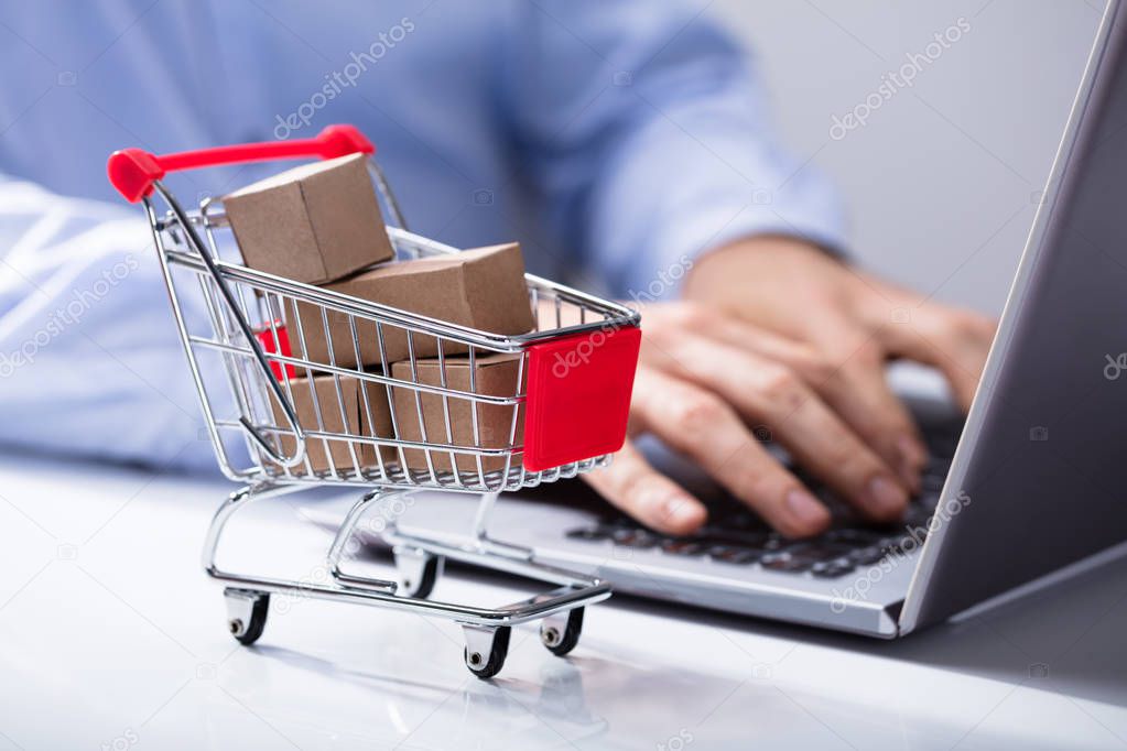 Man Using Laptop With Miniature Cardboard Boxes In The Shopping Trolley On The Desk