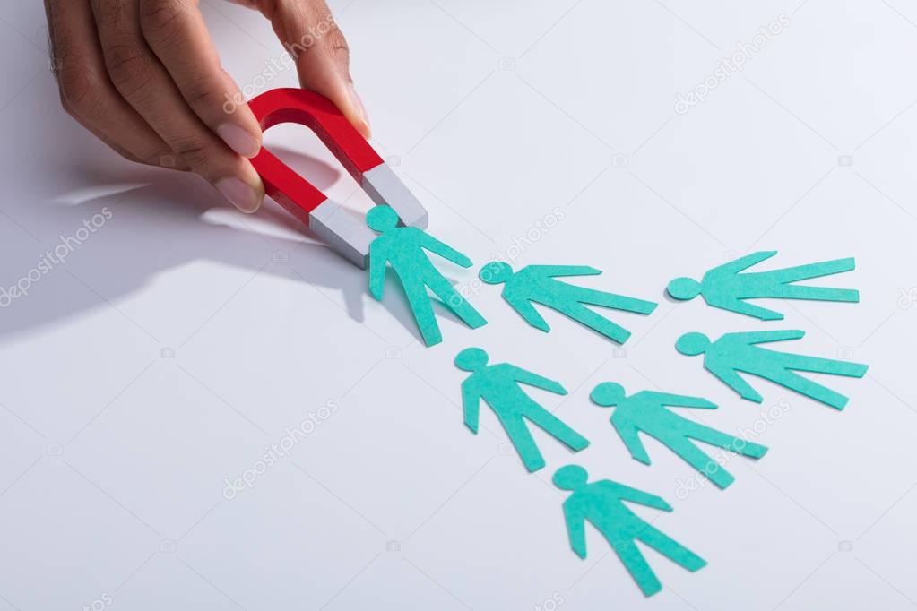 Close-up Of A Businessperson's Hand Holding Red Horseshoe Magnet Attracting Paper Candidates On Desk