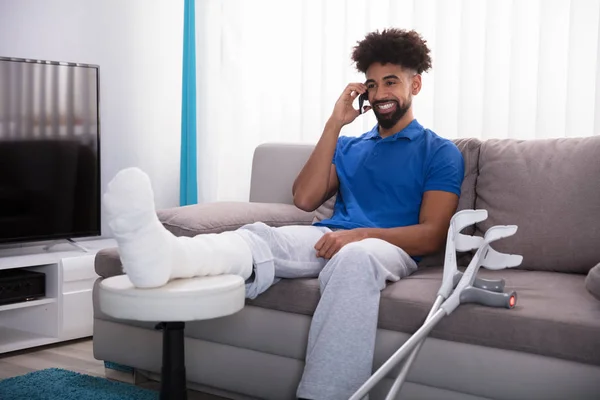 Jovem Feliz Com Perna Quebrada Sentado Sofá Conversando Telefone Móvel — Fotografia de Stock