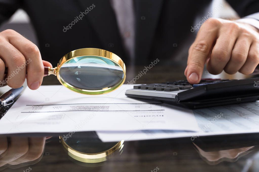 Close-up Of Businessperson's Hand Calculating Bill With Magnifying Glass On Desk