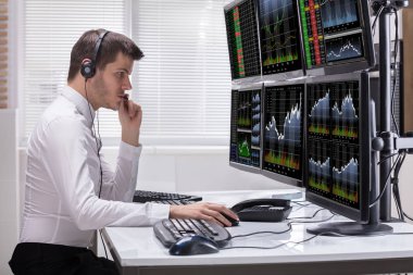 Side View Of A Young Male Stock Market Broker Analyzing Graphs On Multiple Computer Screens