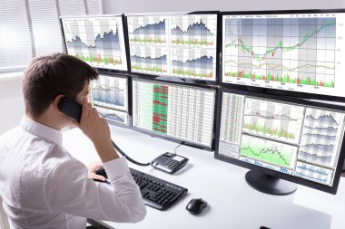 Stok komisyoncu telefon grafikleri gösteren birden çok bilgisayar ekranı önünde konuşurken