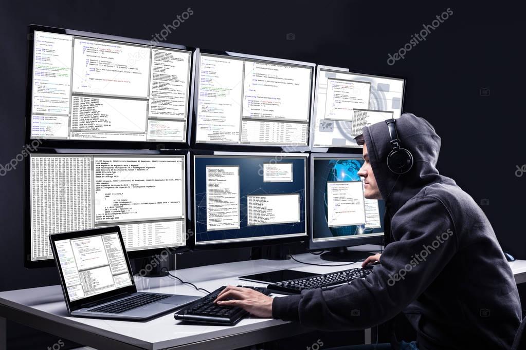 Rear View Of A Hacker Using Multiple Computers For Stealing Data In Office