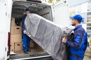 Two Young Delivery Men In Uniform Unloading Furniture From Vehicle clipart