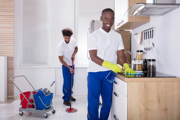 两个微笑的男清洁工在厨房里清洗感应炉和拖把地板 — 图库照片