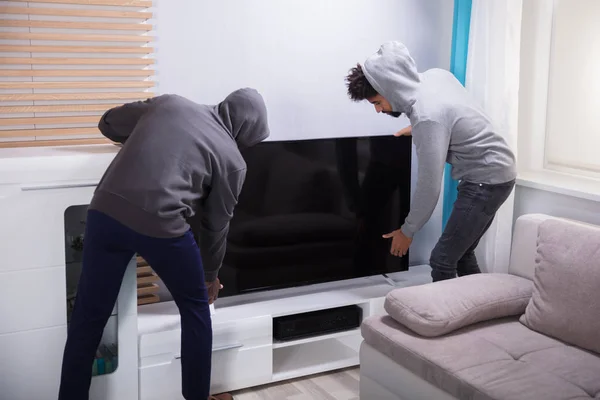 Two Male Robbers In Hooded Top Lifting The Television In The Living Room