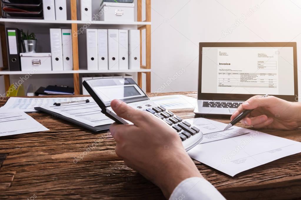 Close-up Of A Businessperson's Hand Calculating Bill With Laptop On Wooden Desk