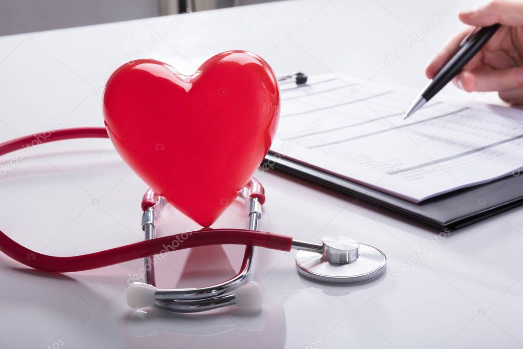 Stethoscope And Red Heart Near Doctor's Hand Analyzing Report On Clipboard