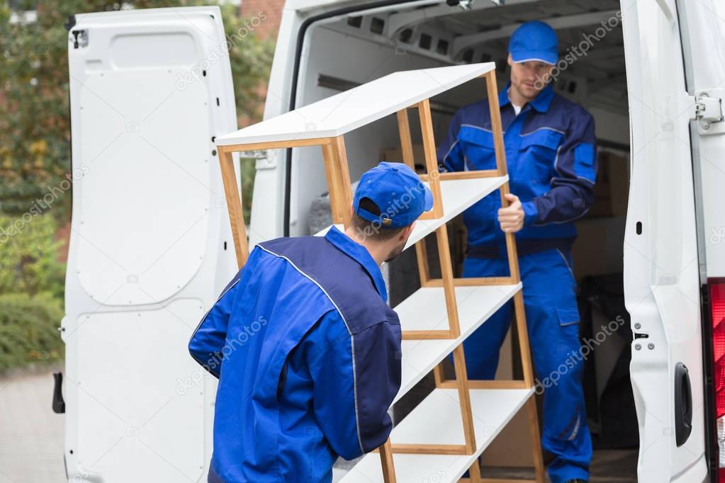 Two Young Delivery Men In Uniform Unloading Wooden Shelf From Truck
