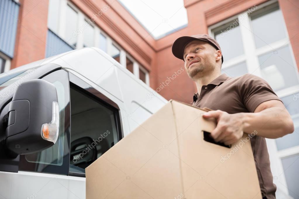 Portrait Of A Happy Young Delivery Man Carrying Cardboard Box Near Van