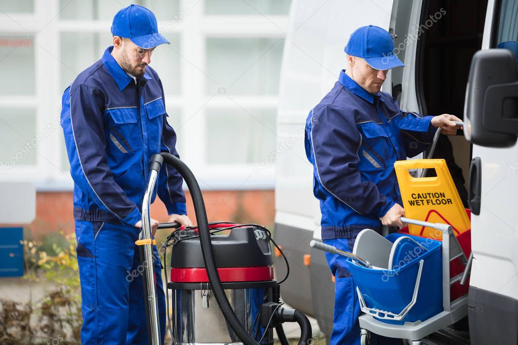Two Young Male Janitor In Blue Uniform Unloading Cleaning Equipment From Vehicle