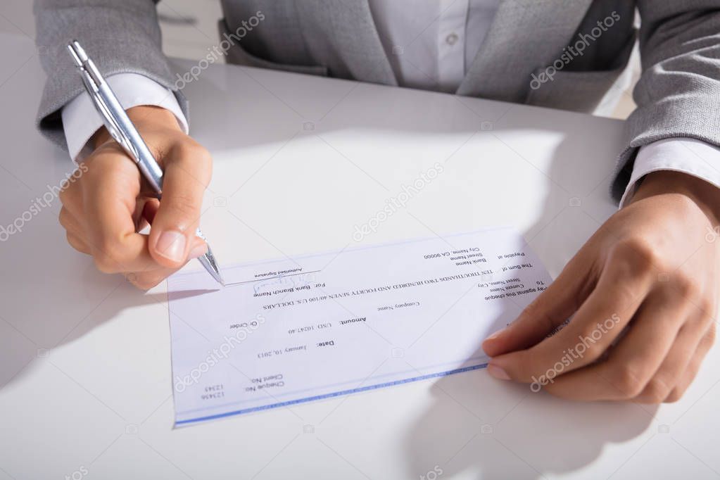 Close-up Of Person Hand Signing Cheque With Pen At Desk