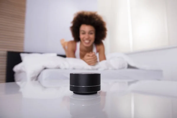 Smiling Young Woman Lying On Bed Listening To Music On Wireless Speaker